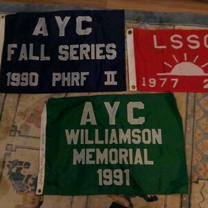 May include: Three rectangular flags with white text on a patterned background. The flags are navy blue, red, and green. The navy flag reads "AYC FALL SERIES 1990 PHRF II". The red flag reads "LSSC 1977 2ND". The green flag reads "AYC WILLIAMSON MEMORIAL 1991".