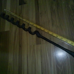 May include: A black auger bit with a curved cutting edge and a long handle, next to a yellow wooden ruler. The auger bit is designed for drilling holes in wood. The ruler measures up to 21 inches.