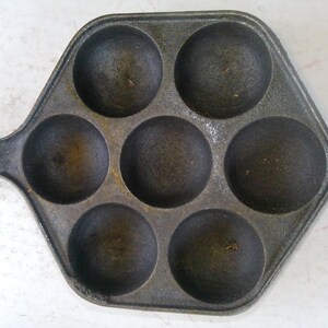 Może przedstawiać: Patelnia z żeliwa w ciemnoszarym kolorze z siedmioma okrągłymi wgłębieniami. Patelnia ma sześciokątny kształt z uchwytem. Jest przeznaczona do gotowania i wydaje się dobrze wypieczona, idealna do naleśnik&oacute;w.