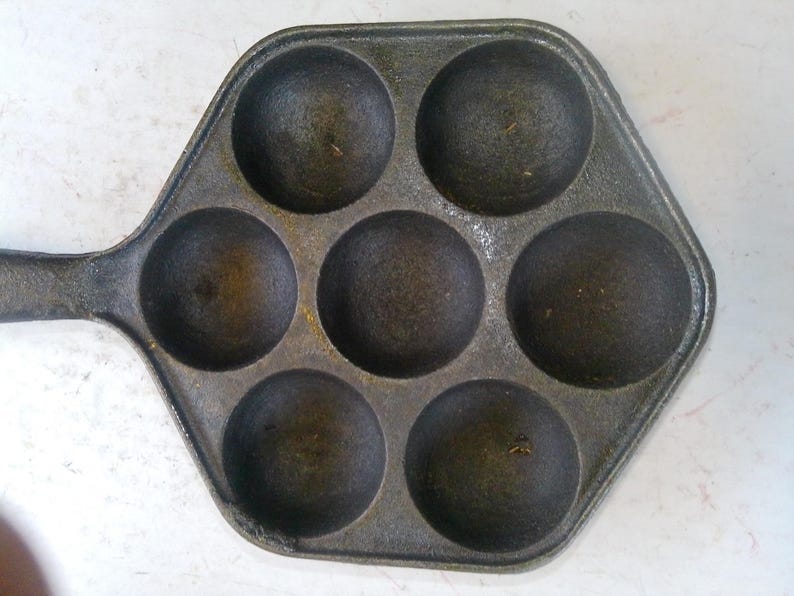 Może przedstawiać: Patelnia z żeliwa w ciemnoszarym kolorze z siedmioma okrągłymi wgłębieniami. Patelnia ma sześciokątny kształt z uchwytem. Jest przeznaczona do gotowania i wydaje się dobrze wypieczona, idealna do naleśnik&oacute;w.