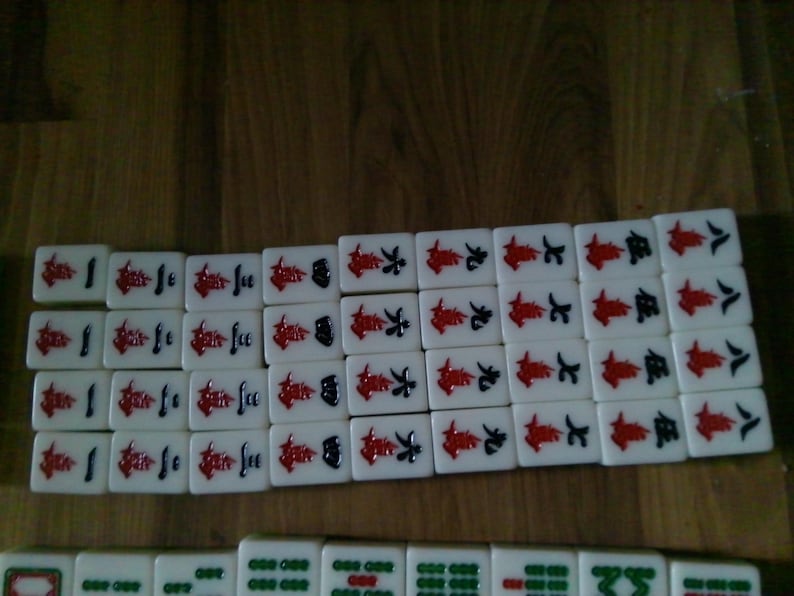 Pu&ograve; includere: Un set di tessere rettangolari da mahjong disposte in file. Le file superiori presentano tessere bianche con caratteri cinesi rossi e neri che rappresentano i numeri da uno a otto. La fila inferiore mostra tessere bianche con motivi circolari verdi.