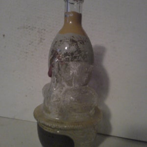May include: A clear glass bottle with a textured surface and a gold top. The bottle is shaped like a person's head and has a black label on the bottom.