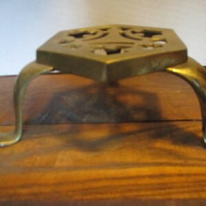 May include: A brass trivet with a hexagonal top and three curved legs. The top features decorative cutouts. The trivet is designed to protect surfaces from hot cookware. The brass has a warm, golden color.