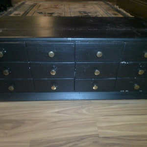 May include: A black wooden chest with 12 drawers, each with a brass knob. The chest is sitting on a wooden floor.