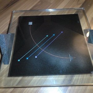 May include: A black square with a copper colored curved line and three blue lines with blue and purple squares at the end of each line. The square is framed in a clear glass frame with black foam padding on the corners.