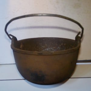 May include: A round, dark brown cast iron pot with a weathered, textured surface and a curved metal handle attached to two side lugs. The vessel features a wide, open top and a deep, rounded base, showing signs of age and oxidation throughout.