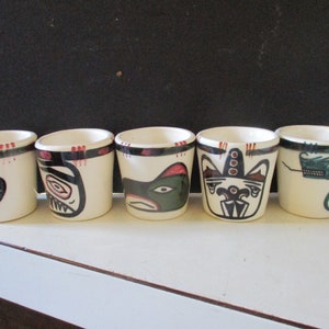 May include: Five white ceramic shot glasses with black, red, and green Native American-inspired designs. Each glass features a different stylized animal or figure. The rims are accented with black and red details.