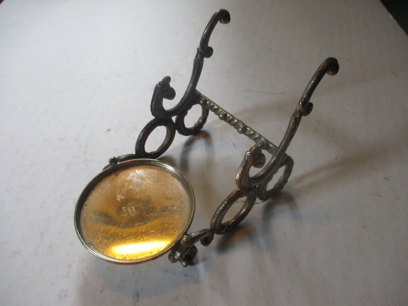 Op de afbeelding: Een gouden metalen standaard met een ronde bovenkant en sierlijke kruldetails. De standaard is ontworpen om een klein object te houden, zoals een fles of een vaas.