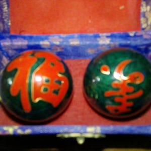 May include: Two spherical metal balls with a dark green enamel finish and bright red Chinese characters, resting inside a rectangular blue fabric-covered box with a red interior lining. The balls feature distinct calligraphic symbols.