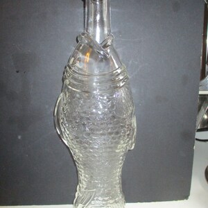 May include: A clear glass bottle shaped like a fish with a textured scale pattern covering the body. The bottle features a narrow neck, a rounded rim, and molded fins on the sides. The design includes detailed gill and tail markings in the glass.