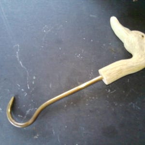 May include: A decorative hook with a duck head handle. The handle is carved from light-colored wood, and the hook is made of brass. The duck head is detailed with a curved neck and beak. The hook is designed for hanging items.