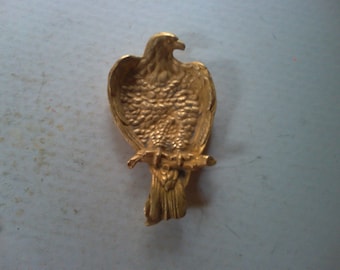 May include: A gold-colored metal eagle figurine with detailed feathers and a perched position. The eagle is facing to the right and has a long, thin beak.
