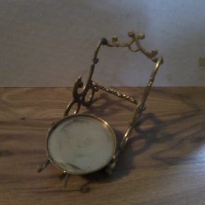 May include: A gold metal stand with a round top and ornate details. The stand is designed to hold a small object, such as a plate or a vase.