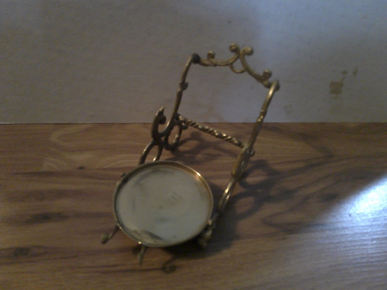 May include: A gold metal stand with a round top and ornate details. The stand is designed to hold a small object, such as a plate or a vase.