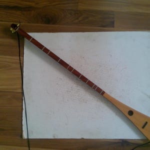 May include: A long, wooden musical instrument with a slender neck and a wide, flat body. The neck is dark brown with white fret markers, and the body is light brown. A black string is attached to the instrument.