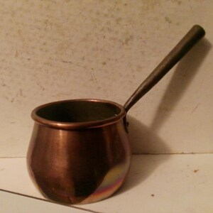 May include: A small, copper-colored pot with a long, dark handle. The pot has a rounded body and a slightly flared rim. The handle is attached to the side of the pot and extends upwards. The pot is likely used for cooking or serving.