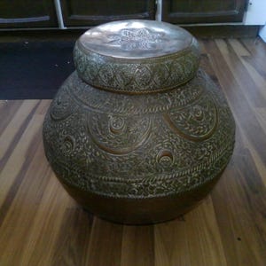 May include: A large, antique bronze-colored metal container with a matching lid. The container and lid are adorned with intricate, embossed floral and geometric patterns. The container is round with a wide base and a smaller, rounded lid.
