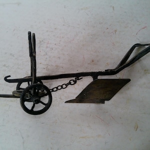 May include: A rustic, miniature metal plow with a dark, aged finish. The plow features a small wheel, a chain, and a blade, evoking a sense of vintage agricultural tools. The design includes handles and a curved plowshare.