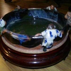 May include: A ceramic bowl with a dark brown glaze and a green interior. The bowl is decorated with small figurines of people in blue and white clothing. The bowl is sitting on a wooden base.