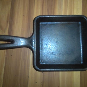 May include: A small, square, cast iron skillet with a long handle. The skillet is black and appears to be well-seasoned. The handle has a hole for hanging. Ideal for individual servings or small portions.