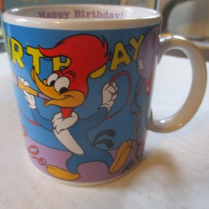 Puede incluir: Una taza de cer&aacute;mica blanca con fondo azul y una imagen de dibujos animados de Woody Woodpecker sosteniendo una bocina de fiesta. La taza tiene las palabras "Happy Birthday!" y "Birthday!" impresas en letras amarillas y rojas.