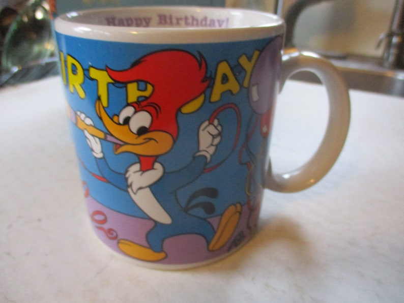 Puede incluir: Una taza de cer&aacute;mica blanca con fondo azul y una imagen de dibujos animados de Woody Woodpecker sosteniendo una bocina de fiesta. La taza tiene las palabras "Happy Birthday!" y "Birthday!" impresas en letras amarillas y rojas.