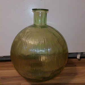 May include: A large, round, olive-green glass vase with vertical ribbed detailing. The vase has a cylindrical neck and a wide, spherical body. The glass has a slight sheen, reflecting light. The vase is a decorative home accent.