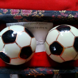 May include: Two decorative soccer ball-shaped objects with black and white panels and copper-colored lines, resting on a red surface inside a patterned box. The box has a black and gold floral design.