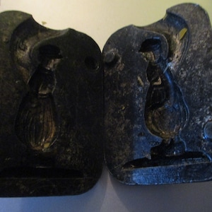May include: Two-part antique metal mold with a woman figure design. The mold is dark gray and rectangular, with a detailed relief of a woman wearing a hat and long dress. The mold is likely for creating decorative items.