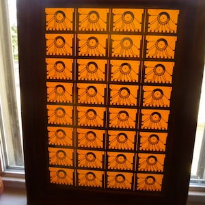 May include: A framed artwork featuring a grid of orange squares, each with a detailed black design. The artwork is encased in a dark brown wooden frame, creating a striking contrast with the bright orange background.