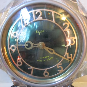 May include: A vintage Majak clock with a faceted, clear plastic frame. The clock face is dark green with gold-colored numerals and hands. The text "Majak" and "11 jewels Made in USSR" are visible.