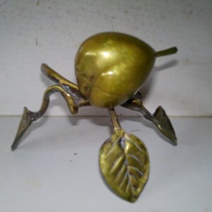 May include: A brass sculpture of a pear with a stem and leaf. The pear is golden and rests on a branch-like base with three pointed legs. The leaf has detailed veining.