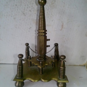 May include: An antique brass stand with a central, spiraled column topped with a ball. The base has a decorative, scalloped edge and is supported by four rounded feet. Small chains connect the four corner posts.