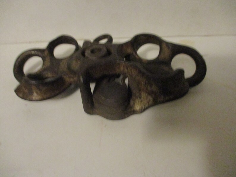 May include: A vintage metal bottle opener with a unique design. The opener has two loops on each side and a central mechanism for opening bottles.