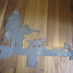 May include: Metal silhouette of a hunter with a gun, a dog, and a tree. The silhouette is mounted on a wooden arrow shape.