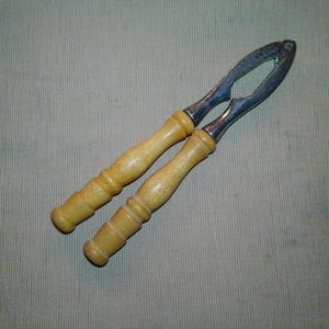 May include: A vintage nutcracker with a metal cracking mechanism and light-colored wooden handles. The nutcracker is designed with a lever-style mechanism for easy use. The handles are smooth and rounded, providing a comfortable grip.