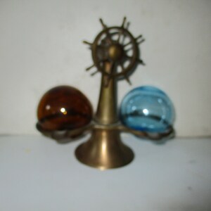 May include: A brass nautical-themed decorative piece featuring a ship's wheel design. It holds two glass spheres, one amber and one blue, in a decorative holder. The piece has a vintage aesthetic.