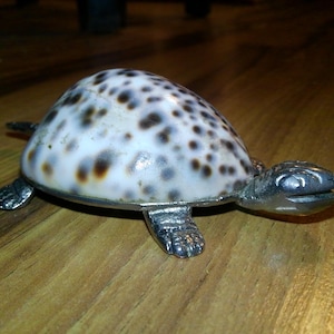 May include: A decorative turtle figurine with a shell patterned with brown spots on a white background. The turtle's head, feet, and trim are silver-colored. The figurine is resting on a wooden surface.