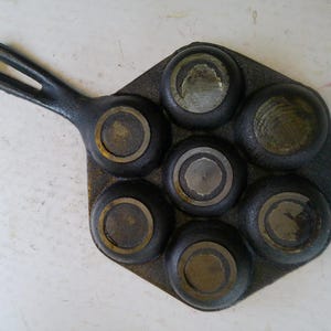 Może przedstawiać: Zabytkowa patelnia do muffin&oacute;w z żeliwa z siedmioma okrągłymi formami. Patelnia ma długi uchwyt i ciemny, postarzany wygląd. Wnętrze każdej formy nosi ślady użytkowania.