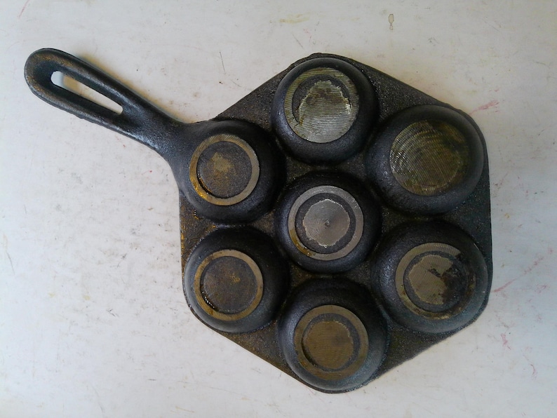 Może przedstawiać: Zabytkowa patelnia do muffin&oacute;w z żeliwa z siedmioma okrągłymi formami. Patelnia ma długi uchwyt i ciemny, postarzany wygląd. Wnętrze każdej formy nosi ślady użytkowania.