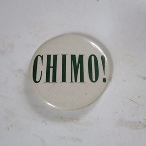 May include: A round, off-white button with the word "CHIMO!" printed in dark green. The button has a glossy finish and is centered in the frame. The background is a textured, off-white surface.
