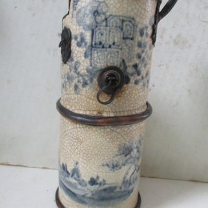 May include: A blue and white ceramic vase with a crackled glaze and metal accents. The vase has a spout and a handle. It is decorated with a landscape scene.