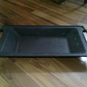 May include: A rectangular, dark gray metal tray with two handles. The tray has a slightly recessed interior and is designed for carrying or serving items. The handles are curved and attached to the sides of the tray.