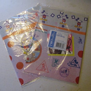 May include: Two sheets of Mickey Mouse gift wrap featuring a blue and pink background with red polka dots. The wrap has a variety of Mickey Mouse images, including Mickey Mouse holding a flag, Mickey Mouse in a circle, and Mickey Mouse in a triangle. The wrap also has a label that says "Disney Gift Wrap" and "Two Big Sheets".