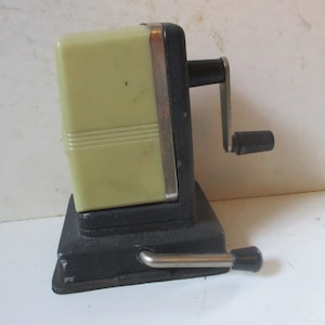 May include: A vintage pencil sharpener with a black base and a pale yellow plastic top. The sharpener has a crank handle and a lever on the side.