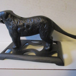 May include: A black cast iron dog-shaped nutcracker. The dog is standing on a rectangular base with decorative cutouts. The nutcracker has a detailed texture and a slot for nuts.