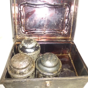 May include: An antique metal tin box with a hinged lid. The box is open and contains three round metal containers with lids. The containers are silver in color and have embossed lettering on the lids.