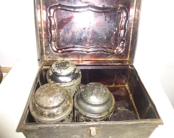 May include: An antique metal tin box with a hinged lid. The box is open and contains three round metal containers with lids. The containers are silver in color and have embossed lettering on the lids.