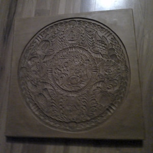 May include: A circular, brown, clay mold with a detailed, intricate design. The design features a central circle with a floral pattern and a surrounding border of repeating motifs.