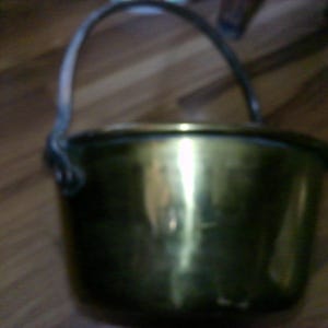 May include: A small, antique brass pot with a curved metal handle. The pot has a rounded body and a dark rim. The brass surface reflects light, creating highlights and shadows. The pot sits on a wooden surface.